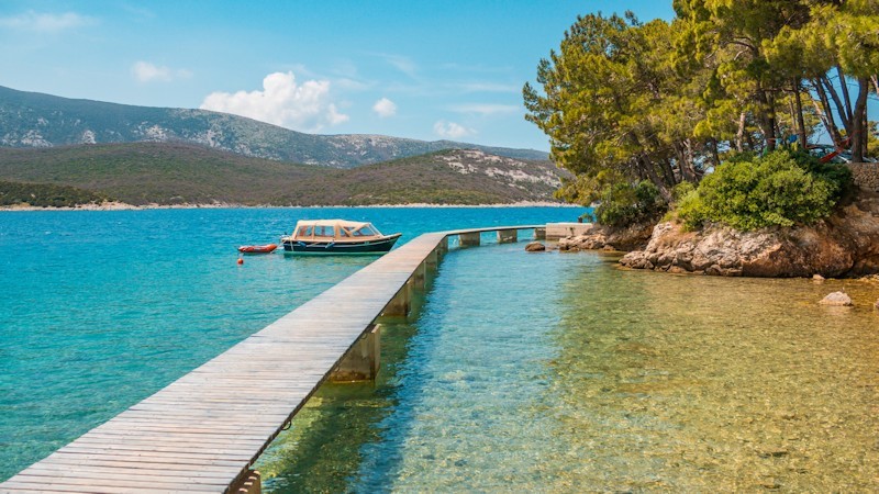 A wooden pier extends into the clear blue sea with a boat nearby and a forested area on the right