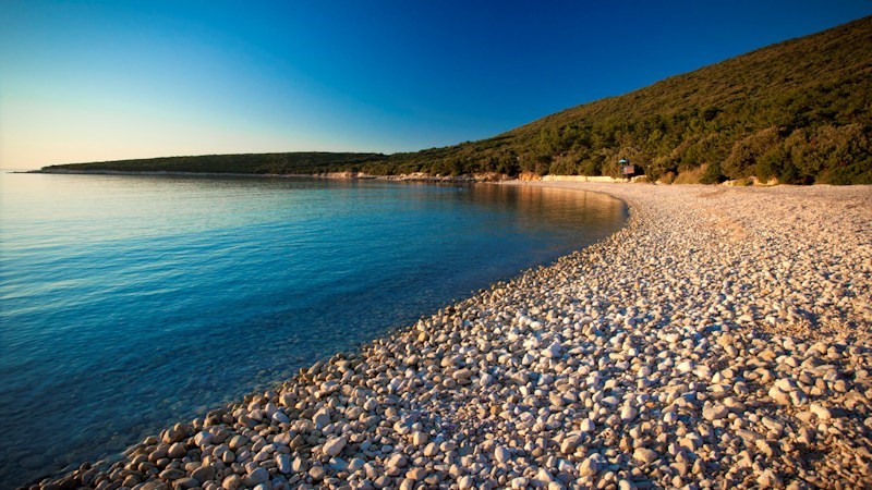Pebble beach on the island of Cres with clear blue sea and a forested hillside